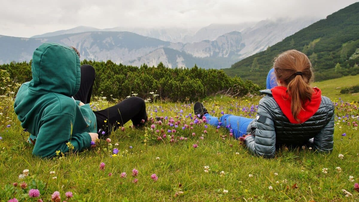 Two people lying on the grass among flowers, facing the mountains.