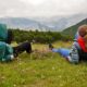Two people lying on the grass among flowers, facing the mountains.
